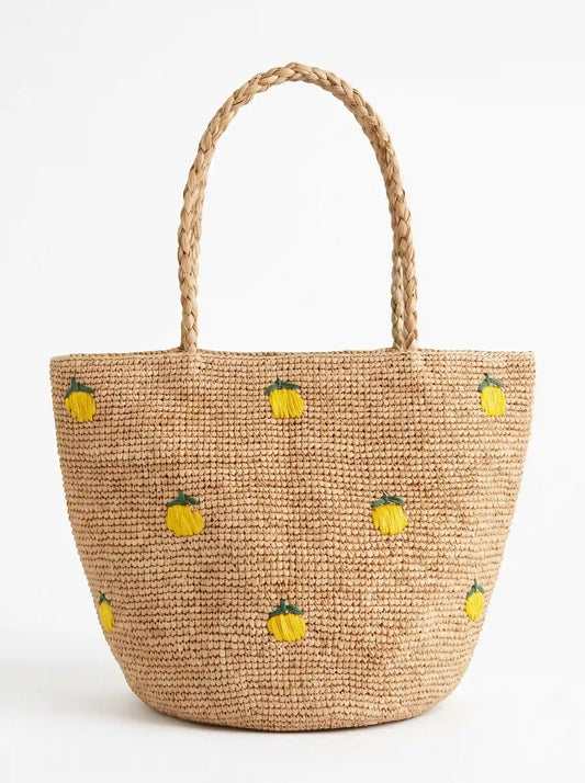 Natural handwoven raffia tote bag with yellow lemon embroidery and braided handles on a white background