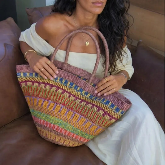 Model holding large multicolor woven raffia tote bag in rainbow stripe pattern, styled with off-shoulder white dress and gold jewelry for resort vacation look