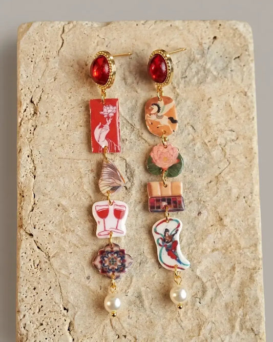 andcrafted asymmetric charm drop earrings with red gemstone studs, illustrated art charms and pearl drops on stone background