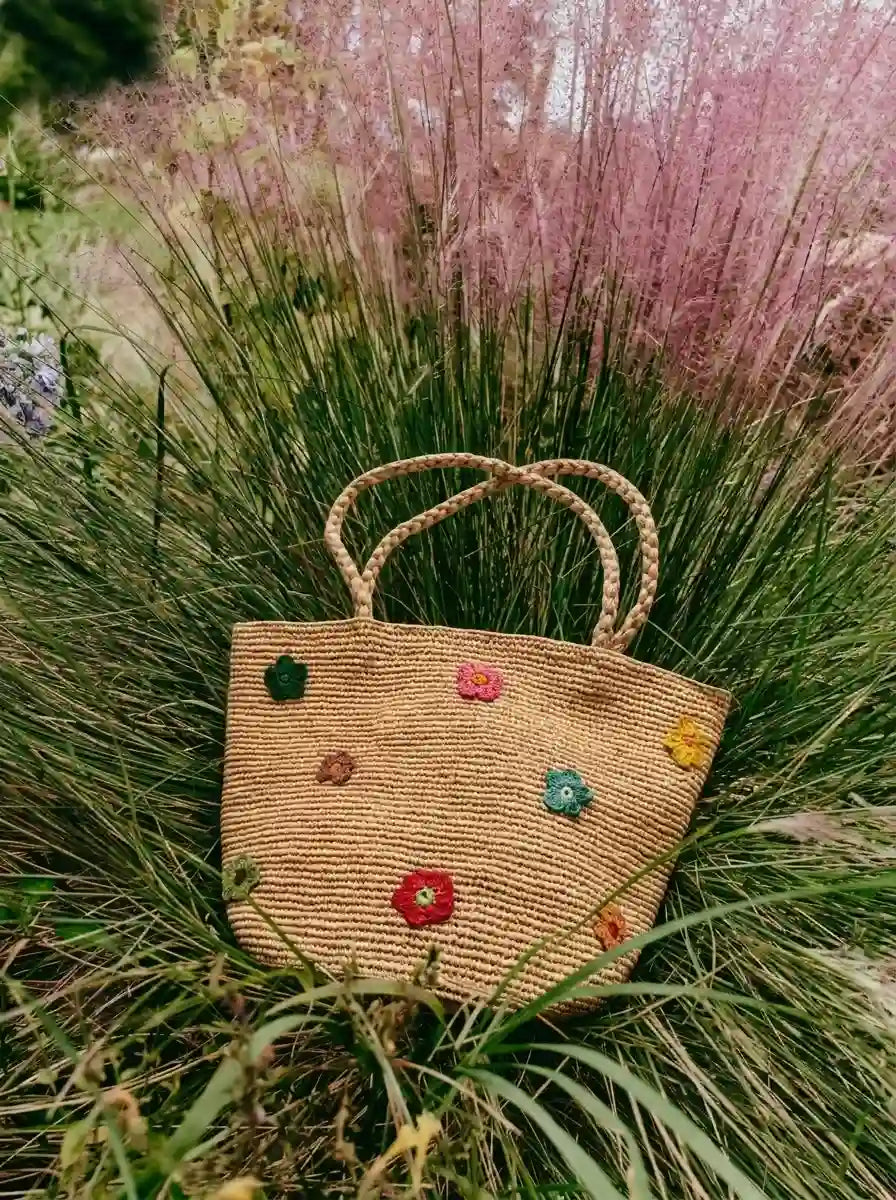 Boho chic handwoven raffia tote bag with colorful crochet flower appliqués placed in lush garden grass