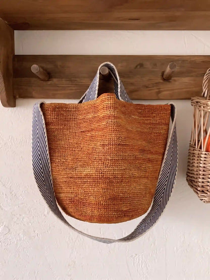 Caramel brown woven raffia tote bag with navy stripe cotton strap hanging on wooden hook rack, cozy slow living home accessories