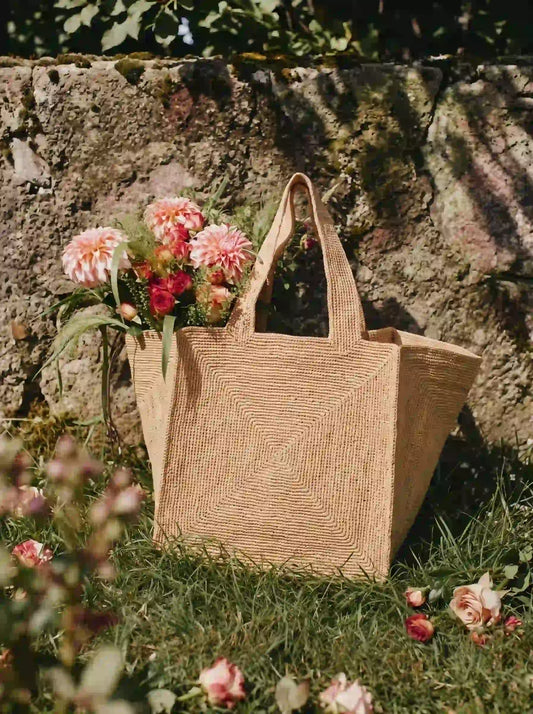 Quiet luxury handwoven raffia tote bag filled with fresh pink dahlias leaning against stone wall in cottage garden