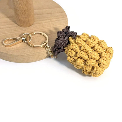  Close-up of a raffia pineapple charm beside a wooden stand, showing chunky yellow stitches, a key ring, and lobster clasp.