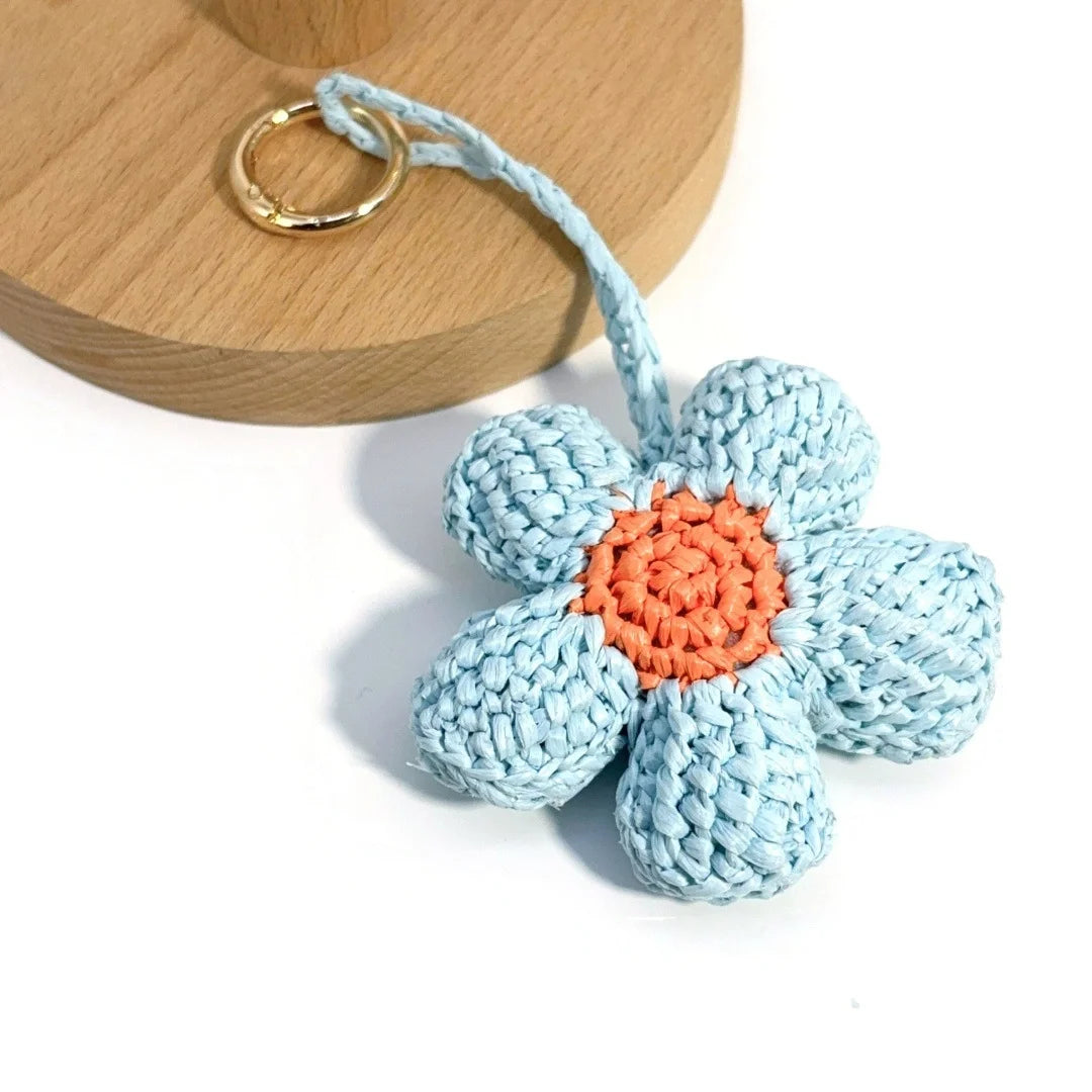 Raffia flower charm with textured blue petals, an orange center, and a gold-tone round clasp beside a wooden display.