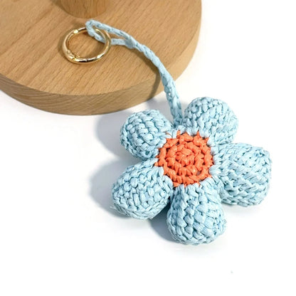 Raffia flower charm with textured blue petals, an orange center, and a gold-tone round clasp beside a wooden display.