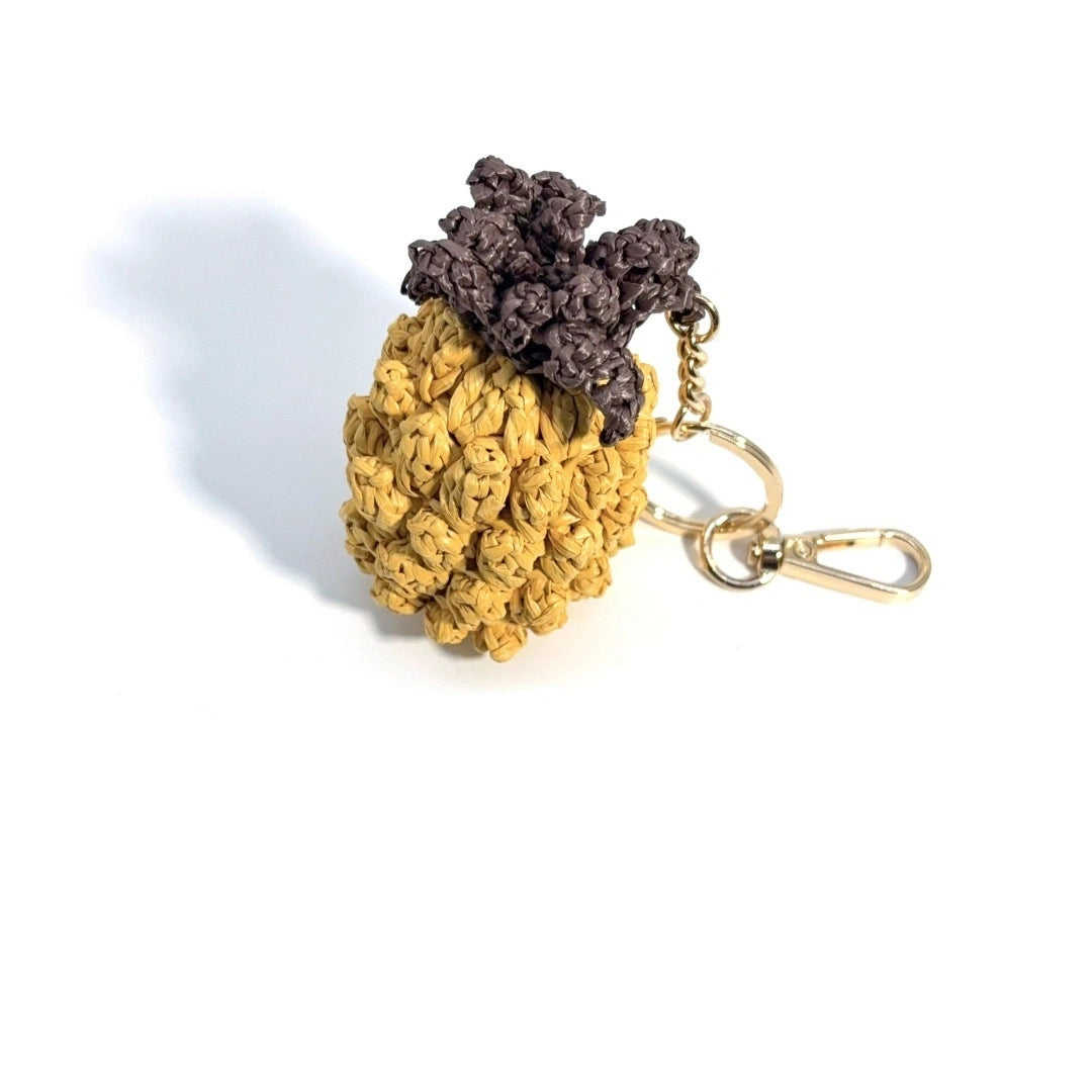 Yellow hand-crocheted raffia pineapple bag charm with a dark brown leafy crown and gold-tone hardware on white.