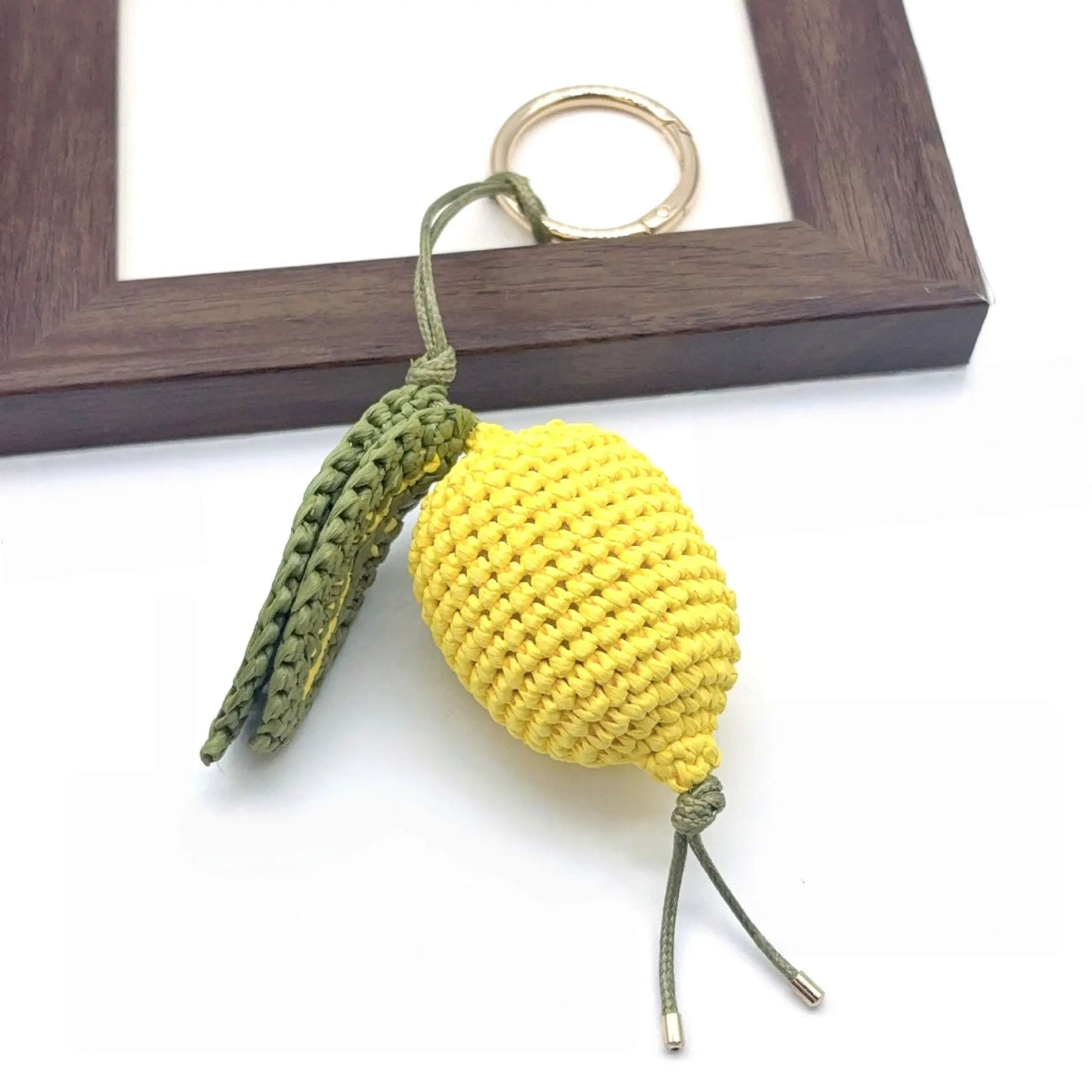 Handmade raffia lemon charm beside a wooden frame, showing crochet texture, green leaves, and cord details.