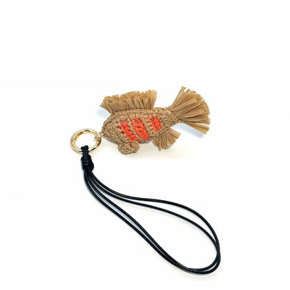 Handmade raffia fish bag charm laid flat with a long black cord loop, gold-tone clasp, and fan-shaped fringed tail.
