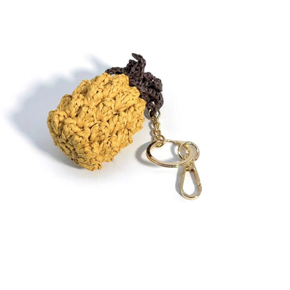 Handmade raffia pineapple bag charm lying on its side, with a textured yellow body, brown crown, and gold-tone clip.