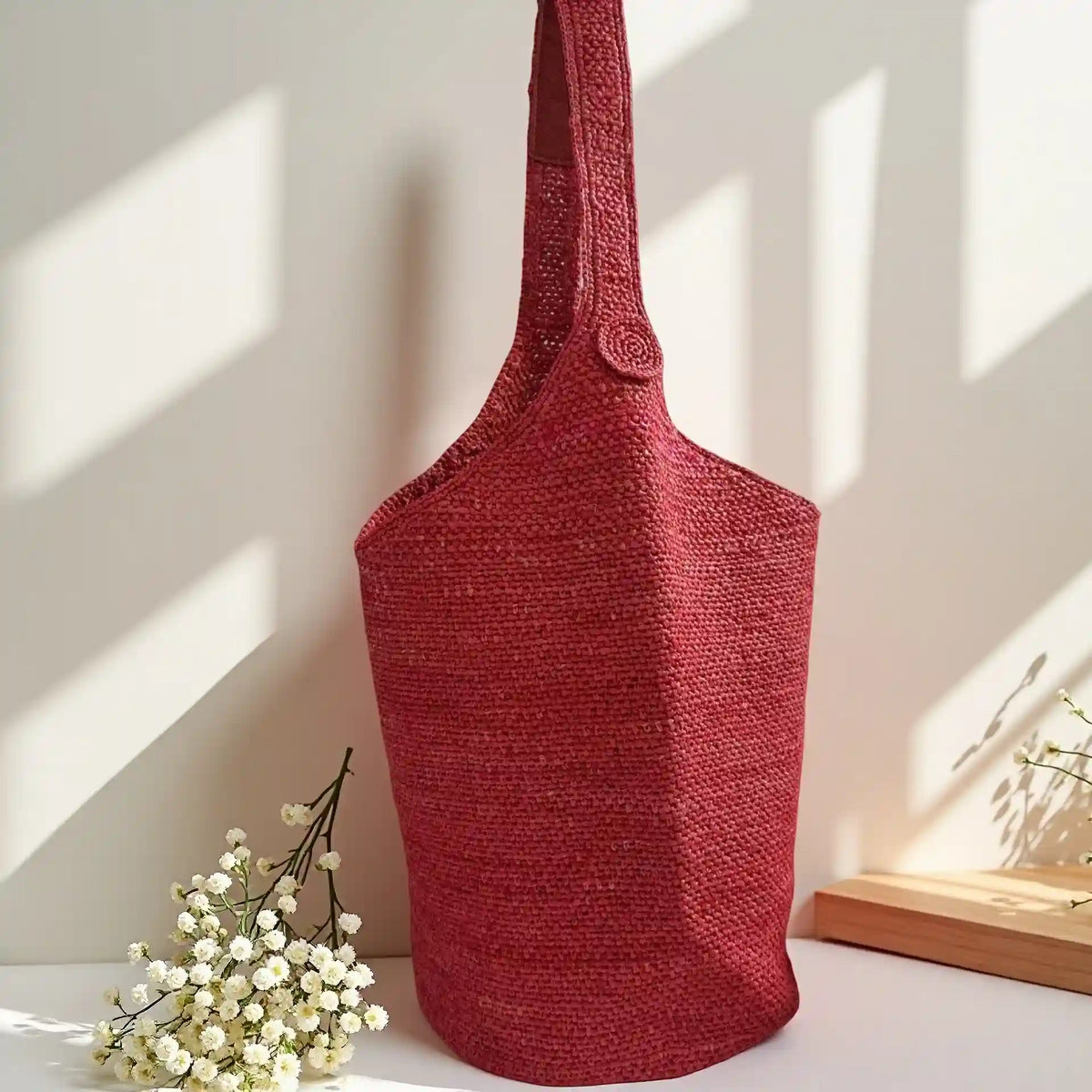 Burgundy red handwoven raffia bucket bag with single shoulder strap and crochet button closure, boho chic woven purse in warm sunlight